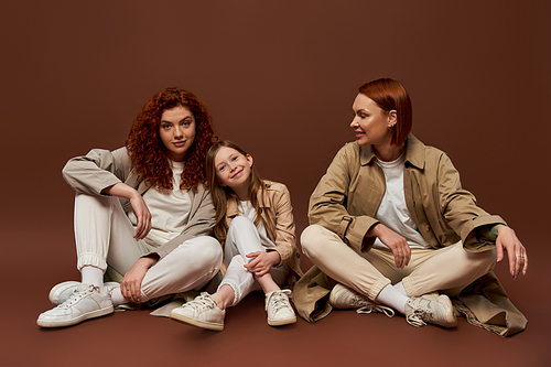 cheerful redhead family of three female generations sitting on brown background, autumn coats
