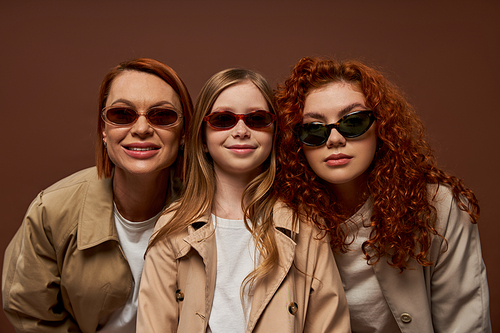 portrait of happy three generations of women looking at camera in sunglasses on brown backdrop