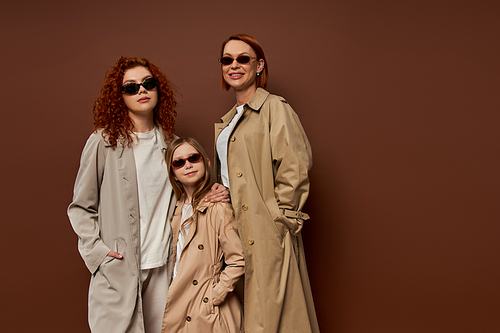family portrait of female generations in sunglasses and coats on brown background, hands in pockets