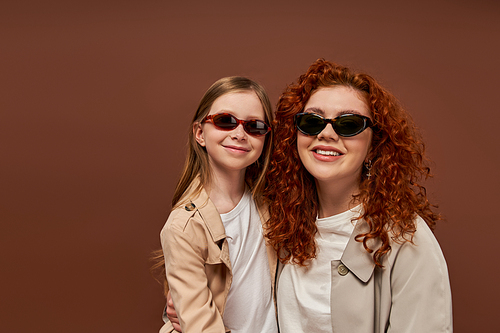 two generations, happy curly mother and daughter in sunglasses on brown backdrop, portrait