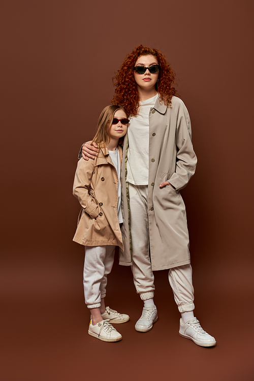 generations, redhead mother and child posing in sunglasses and beige autumnal coats, brown backdrop