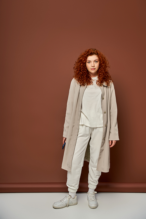 redhead and curly young woman in beige trench coat and joggers standing on brown background