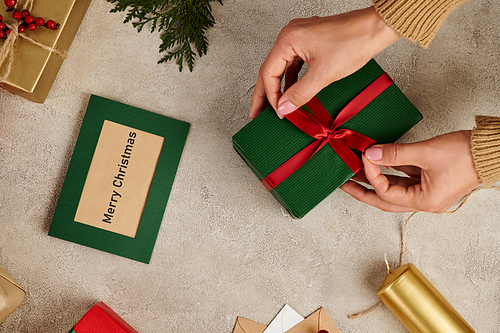 cropped view of woman tying red bow on gift box near Merry Christmas greeting card and festive decor