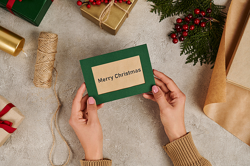 cropped woman with handmade Merry Christmas greeting card near juniper branches and holly berries