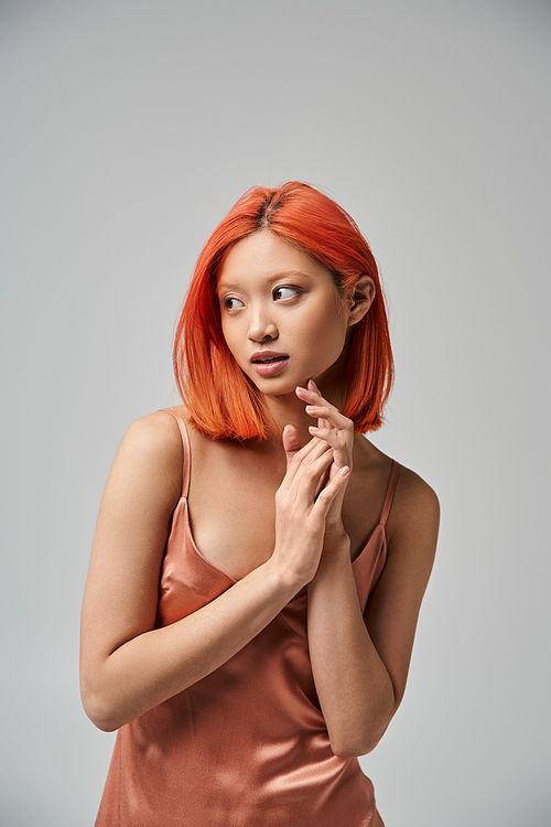 portrait of dreamy young asian woman with red hair posing in silk slip dress on grey background