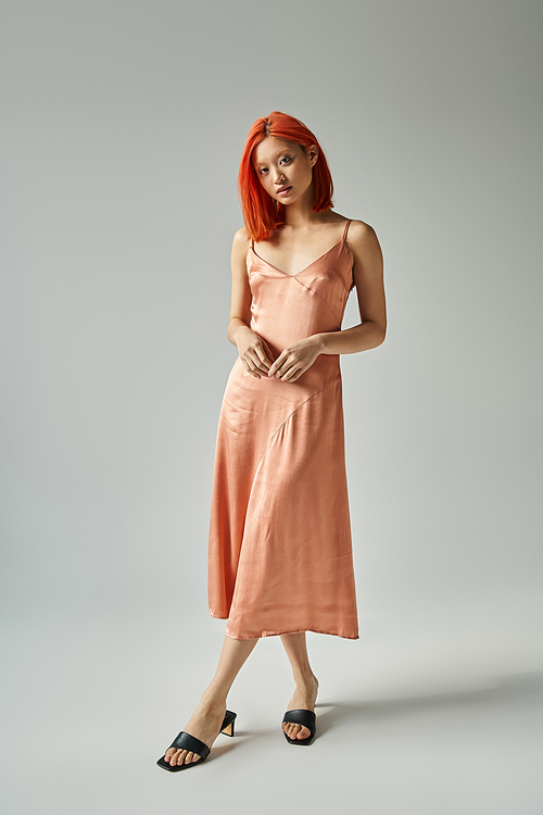 sensual asian woman with red hair posing in silk slip dress on grey background, female elegance