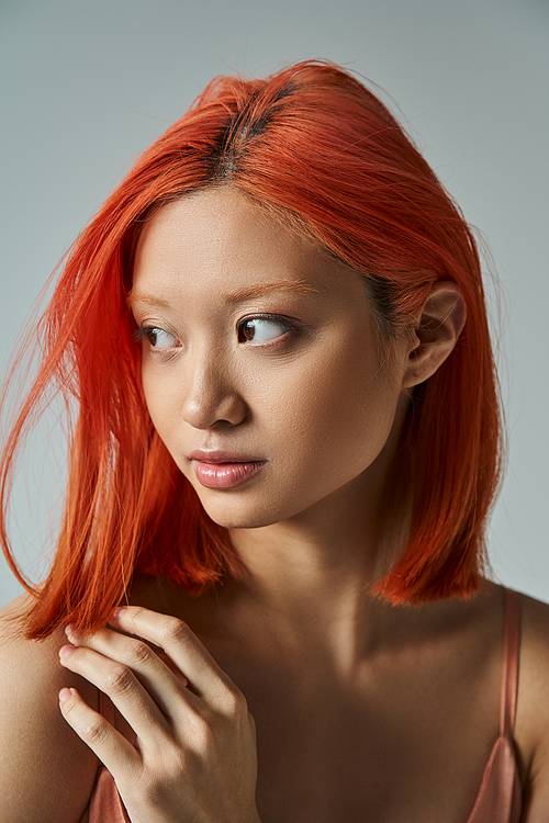 elegant and young asian woman with red hair and natural makeup looking away on grey backdrop