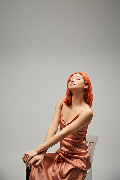 asian woman with closed eyes sitting on chair in silk slip dress on grey background, serenity