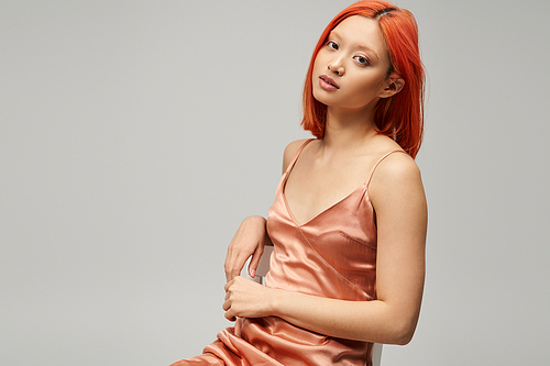 young alluring asian woman sitting on chair in silk slip dress on grey background, relaxed pose