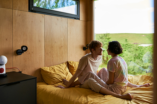 multiethnic couple looking at each other and sitting on bed in country house with forest view