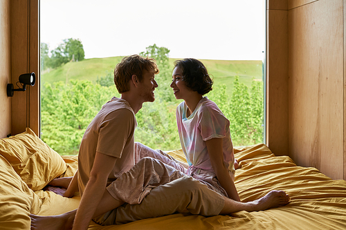 joyful multiethnic couple looking at each other and sitting on bed in country house with forest view