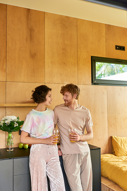 happy multicultural couple standing in pajamas and holding glasses with orange juice in morning