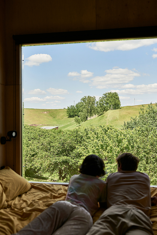 scenic view, back view of couple lying on bed and looking at green trees on hill behind window