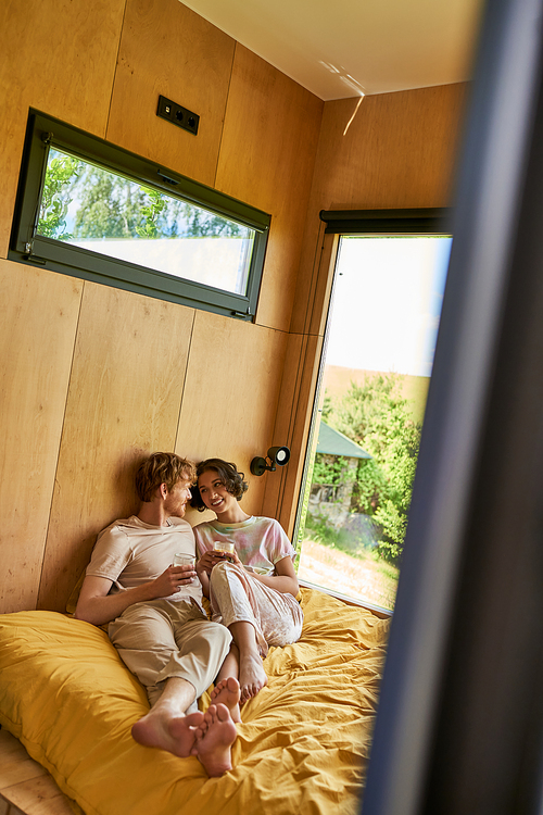 cheerful interracial couple sitting  on bed and holding cups of morning coffee in country house