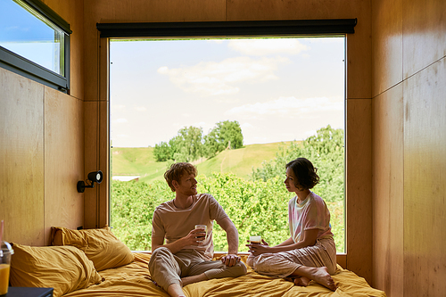 interracial couple holding cups of coffee and sitting on bed next to window with beautiful view