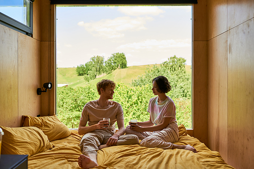 happy diverse couple holding cups of coffee and sitting on bed next to window with beautiful view