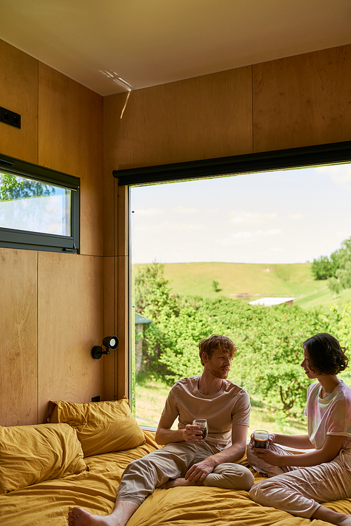 multicultural couple holding cups of coffee and sitting on bed next to window with beautiful view