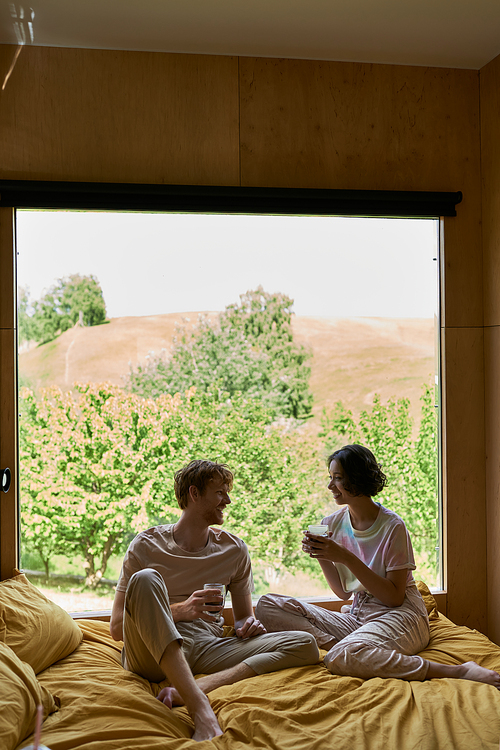 happy multiethnic couple holding cups of coffee and sitting on bed next to window with natural view