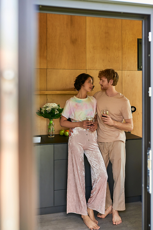 beautiful interracial couple standing in pajamas and holding cups with morning coffee in kitchen