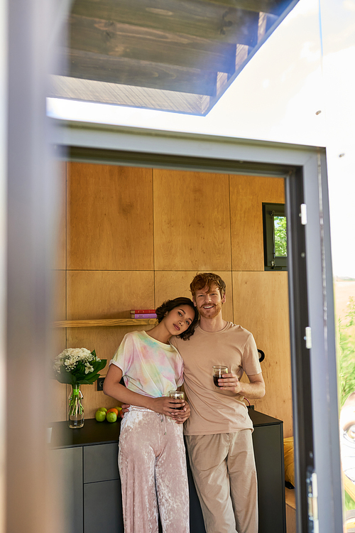 beautiful multiethnic couple standing in pajamas and holding cups with morning coffee in kitchen