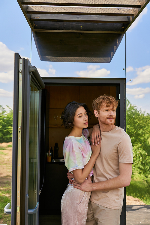 happy interracial couple standing at front door of modern glass house and looking away, fresh air