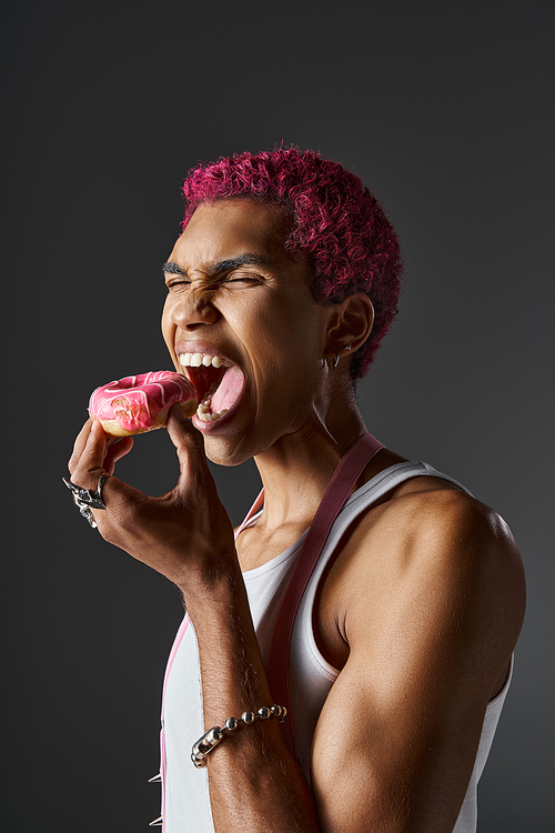 voguish handsome man with pink hair and accessories eating delicious pink donut, fashion and style