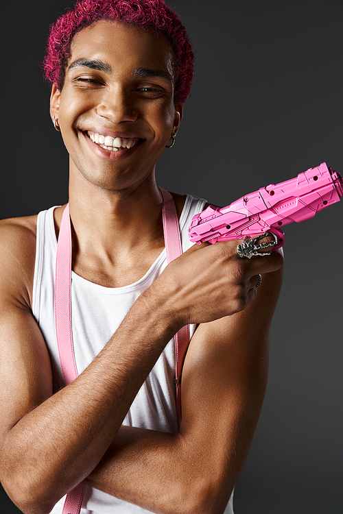 portrait of cheerful young man with pink hair and suspenders posing toy gun and smiling happily