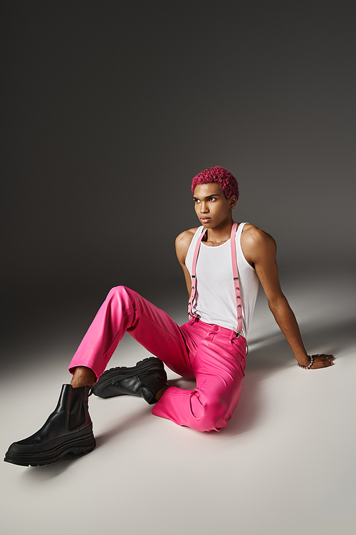 handsome pink haired man in vivid bold attire sitting on floor and looking away, fashion concept