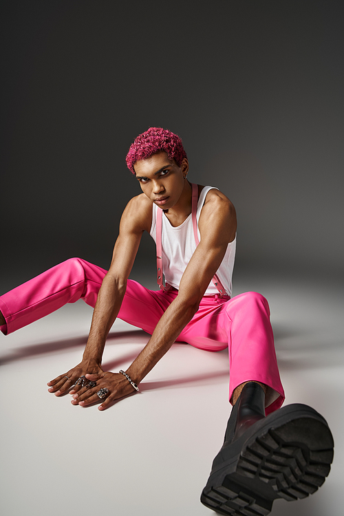 stylish young man in pink pants with suspenders sitting on floor on gray backdrop, fashion concept
