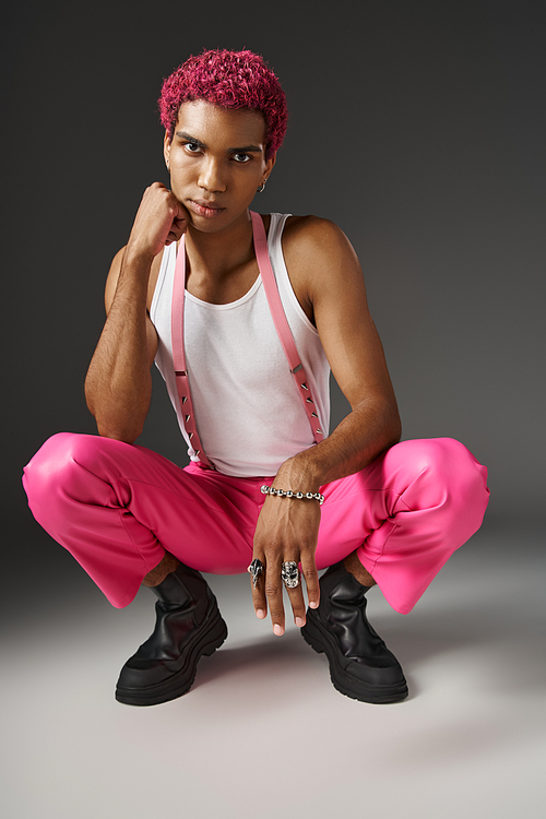 trendy african american man posing on gray backdrop squatting and looking at camera, fashion concept