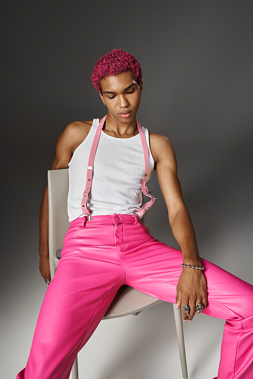 handsome stylish man in pink pants with suspenders posing on chair on gray backdrop, fashion concept