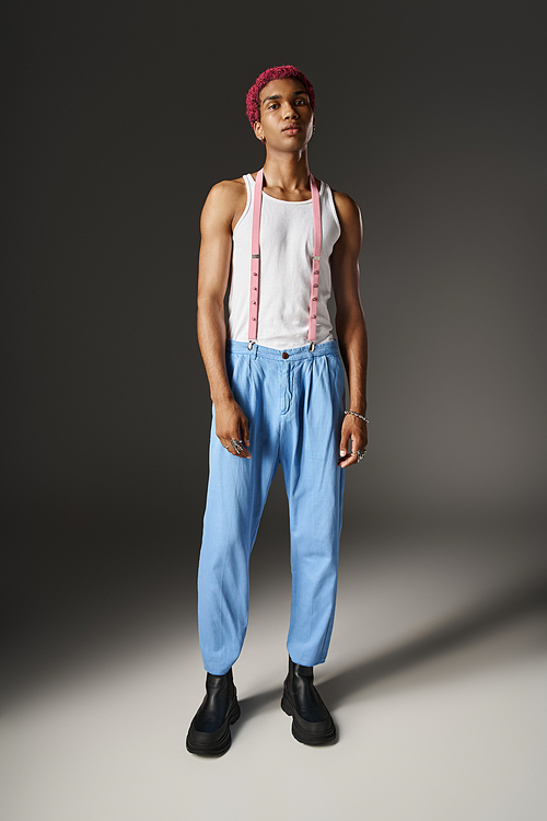 handsome man with pink hair on gray backdrop in blue pants with pink suspenders, fashion concept