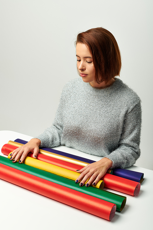 young woman with short hair looking at colorful gift paper on table with grey backdrop, Christmas