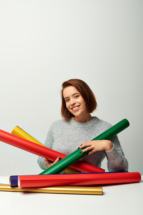 happy woman with short hair holding colorful wrapping paper on table with grey backdrop, Christmas
