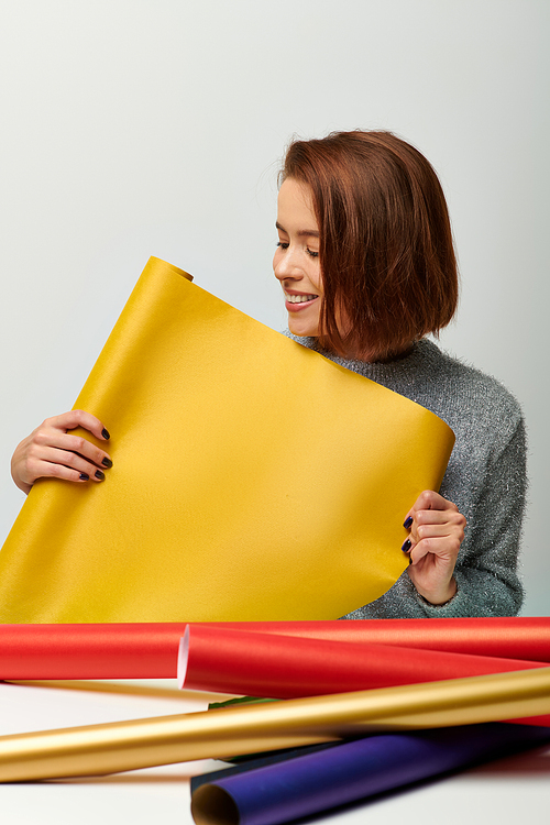 smiling young woman in sweater holding yellow gift paper on grey backdrop, Merry Christmas concept