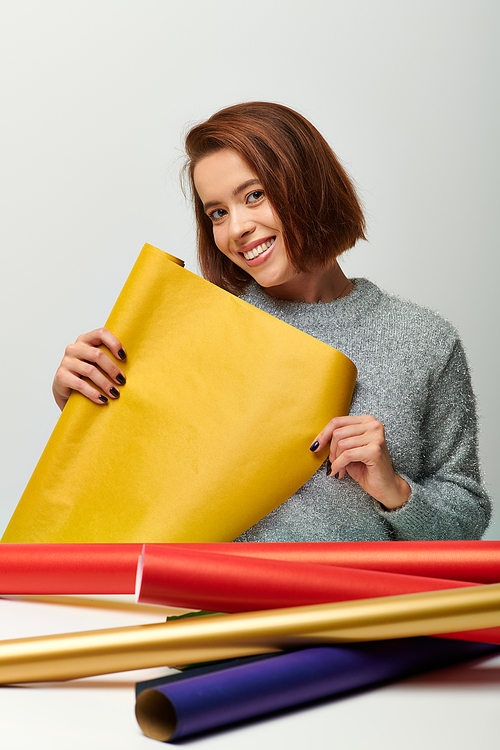 smiling woman in cozy sweater holding yellow gift paper on grey backdrop, Merry Christmas concept