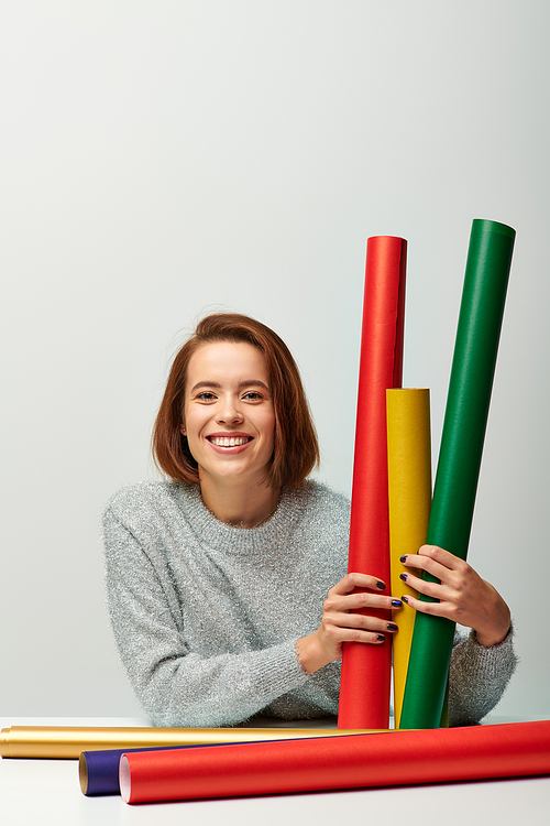 smiling woman in cozy sweater holding different colors gift paper on grey backdrop, Merry Christmas