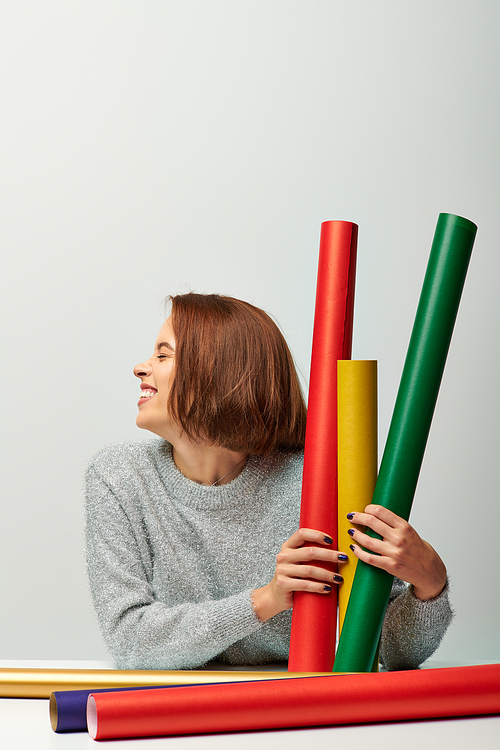 joyous woman in cozy sweater holding different colors gift paper on grey backdrop, Merry Christmas