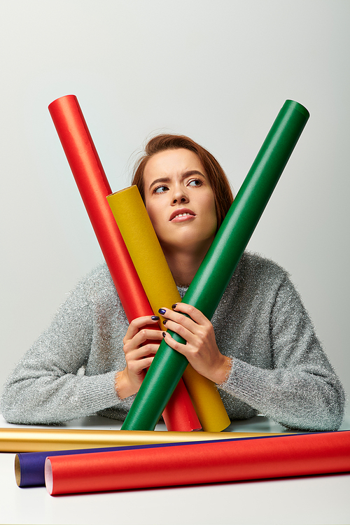 pensive woman in winter sweater holding colorful wrapping paper on grey backdrop, Merry Christmas