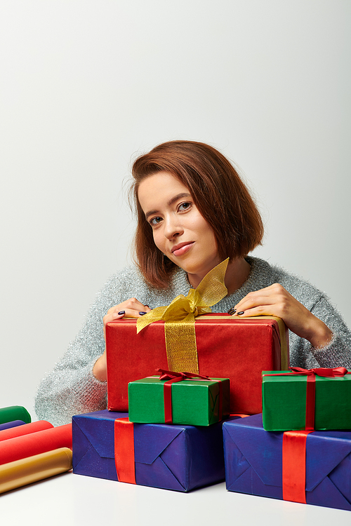 young woman in winter sweater holding wrapped present near colorful gift paper on grey, Christmas