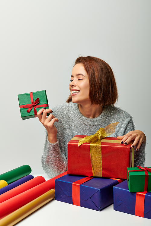 happy woman in winter sweater looking at wrapped Christmas present near colorful gift paper on grey