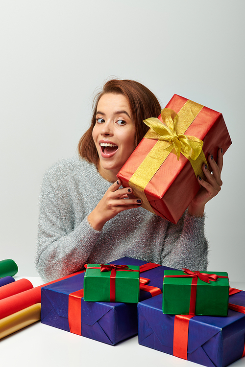 happy woman in winter sweater holding red Christmas present near gift paper on grey backdrop