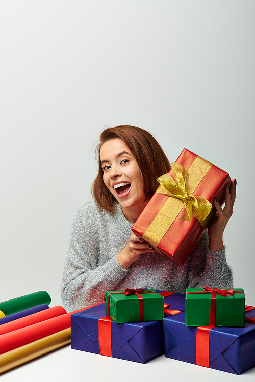 thrilled woman in winter sweater holding red Christmas present near gift paper on grey backdrop