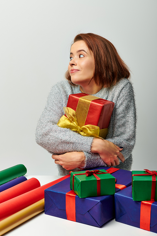 thrilled woman in winter sweater hugging red Christmas present near gift paper on grey backdrop