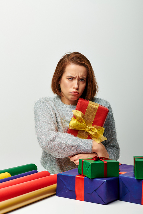 upset woman in winter sweater holding red wrapped Christmas present near gift paper on grey backdrop