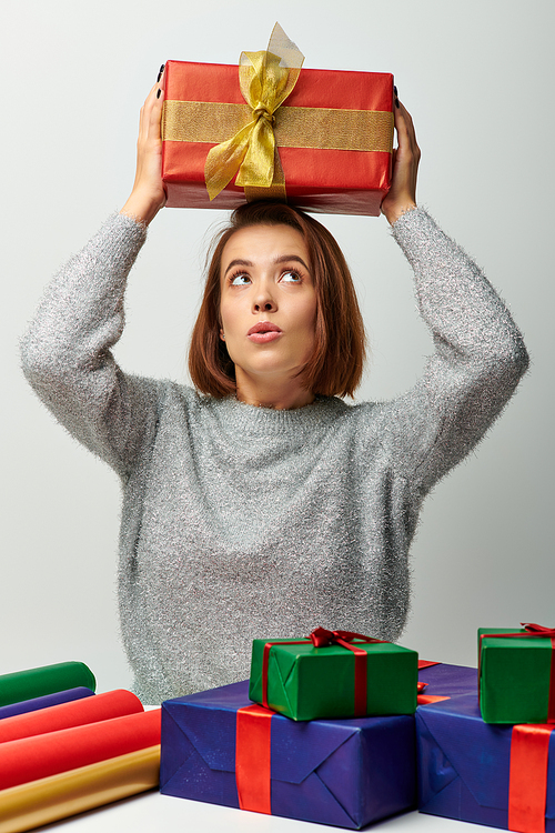 thrilled woman in winter sweater holding Christmas present above head near gift paper on grey