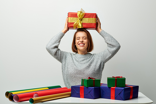 cheerful woman in winter sweater holding Christmas present above head near gift paper on grey