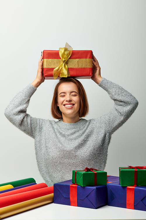 pleased woman in winter sweater holding Christmas present above head near gift paper on grey