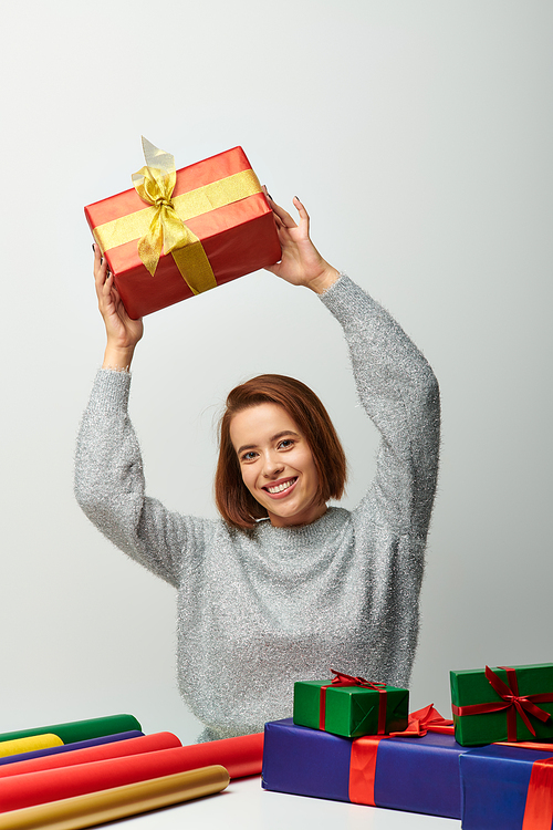 smiling woman in winter sweater holding Christmas present above head near gift paper on grey