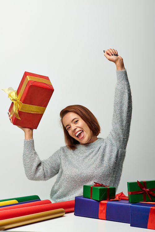 enthusiastic woman in winter sweater holding Christmas present above head near gift paper on grey